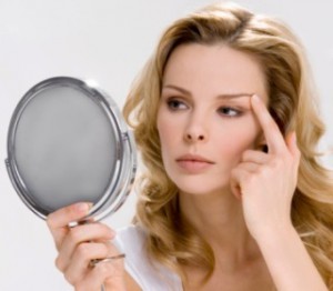 Young woman looking at reflection in hand mirror, touching eyebrow